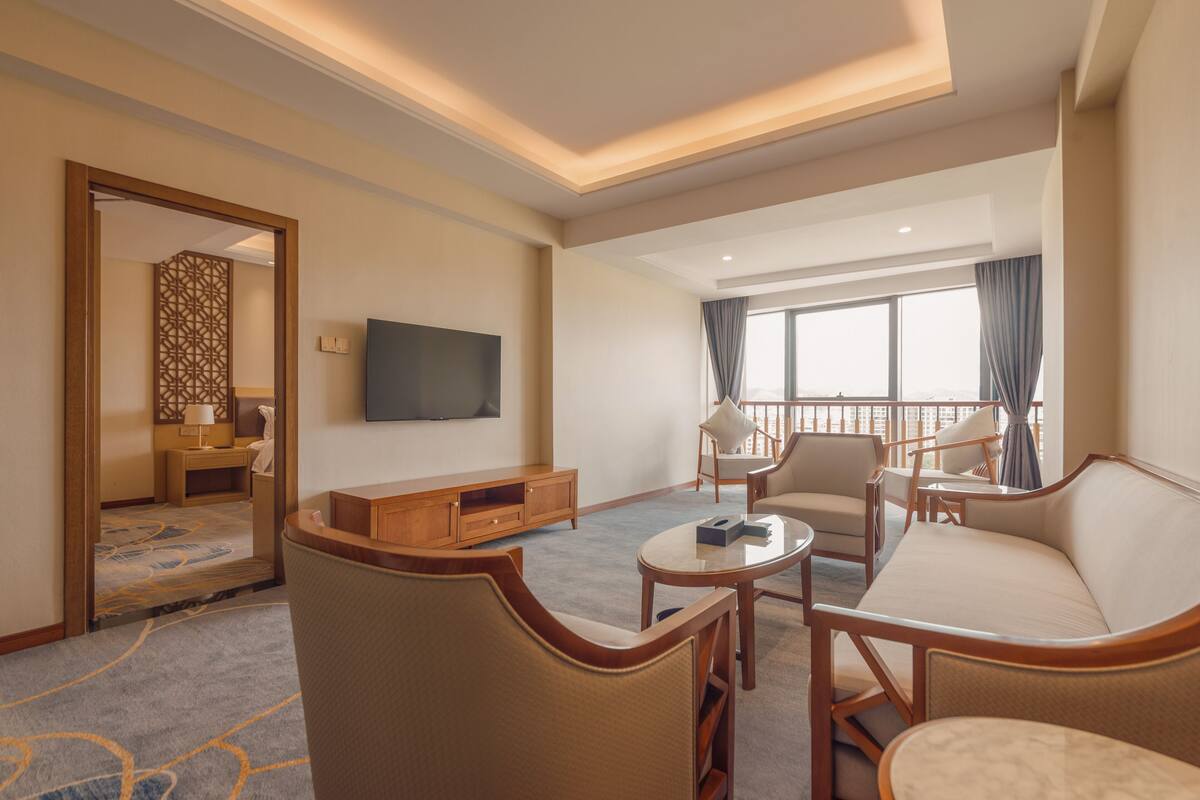 Habitación de hotel con sala de estar que incluye un sofá, sillas, una mesa de centro, televisor y un balcón con vistas.