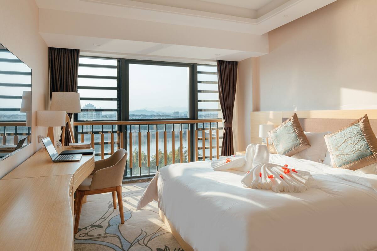 Habitación de hotel con una cama grande, un escritorio con una computadora portátil y vistas de la ciudad a través de amplias ventanas.