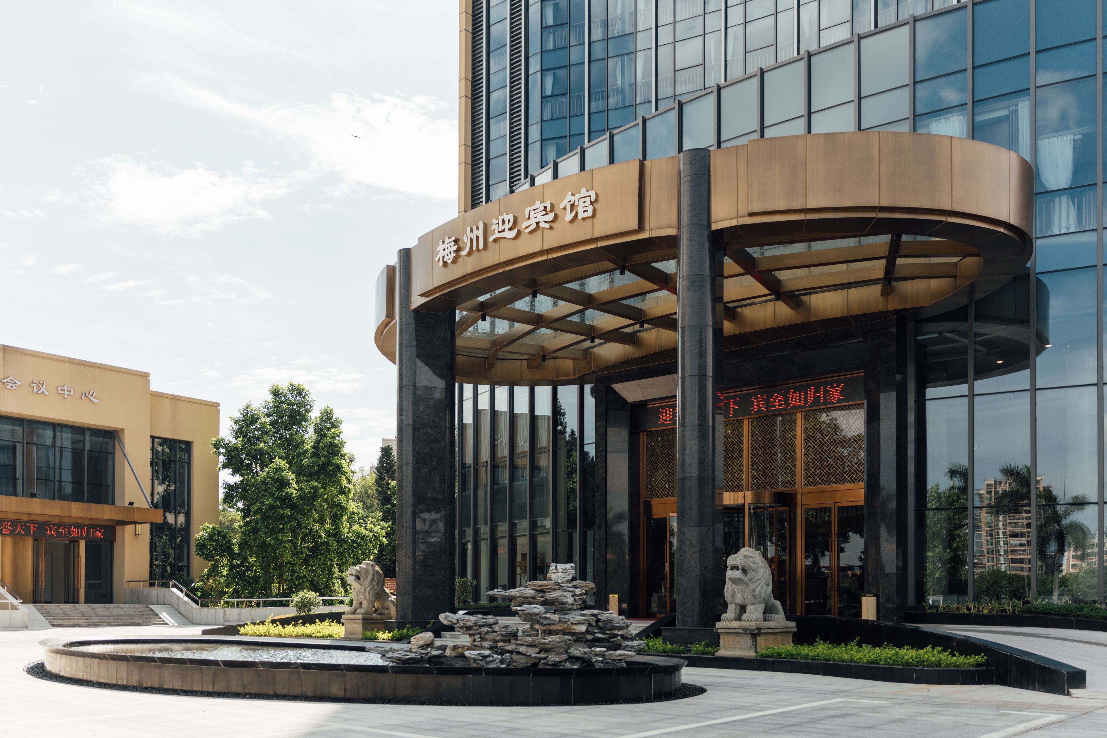 A modern hotel entrance with a curved canopy, a stone fountain, and two lion statues.