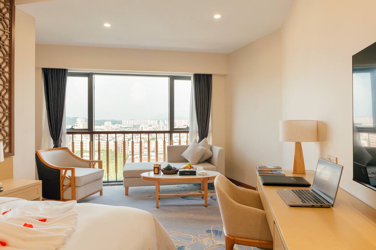 Habitación de hotel con cama, dos sillones, un sofá, una mesa de centro, un escritorio con una computadora portátil, una lámpara y un televisor.