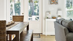 Una habitación con una mesa de comedor de madera, sillas de mimbre, un sofá gris, una pequeña mesa blanca con una lámpara y una puerta que da a un balcón con árboles.