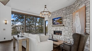 A modern living room with a stone fireplace, a white sofa, a wooden coffee table, and a large window offering a view of trees and mountains.