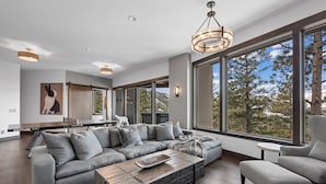 A modern living room with a large gray sectional sofa, a wooden coffee table, and a gray armchair. There is a large window offering a view of trees and mountains.