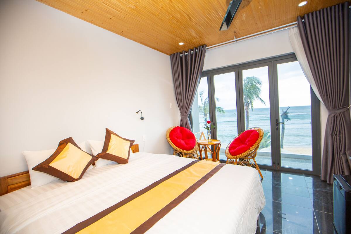 Une chambre d’hôtel avec un grand lit, deux fauteuils rouges, une petite table et une vue sur l’océan.
