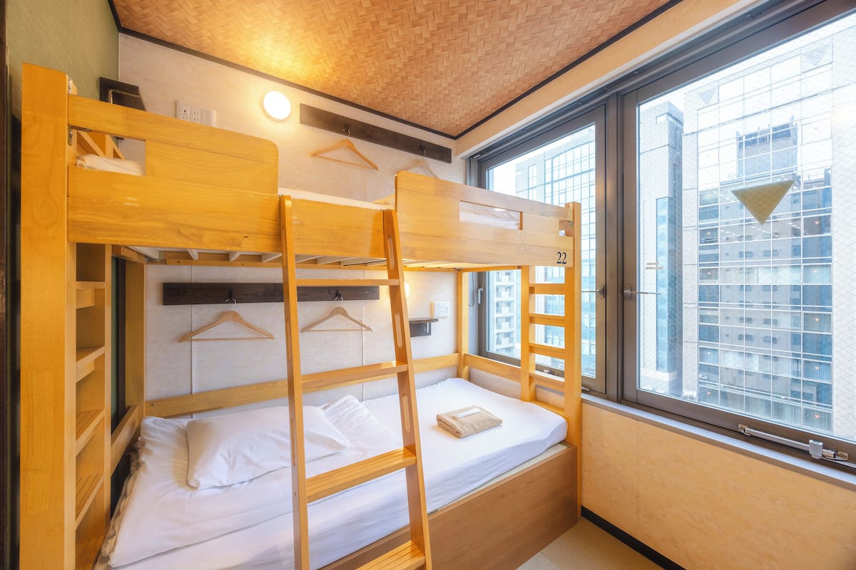 Une chambre avec un lit superposé en bois, une fenêtre donnant sur des immeubles et un luminaire suspendu.