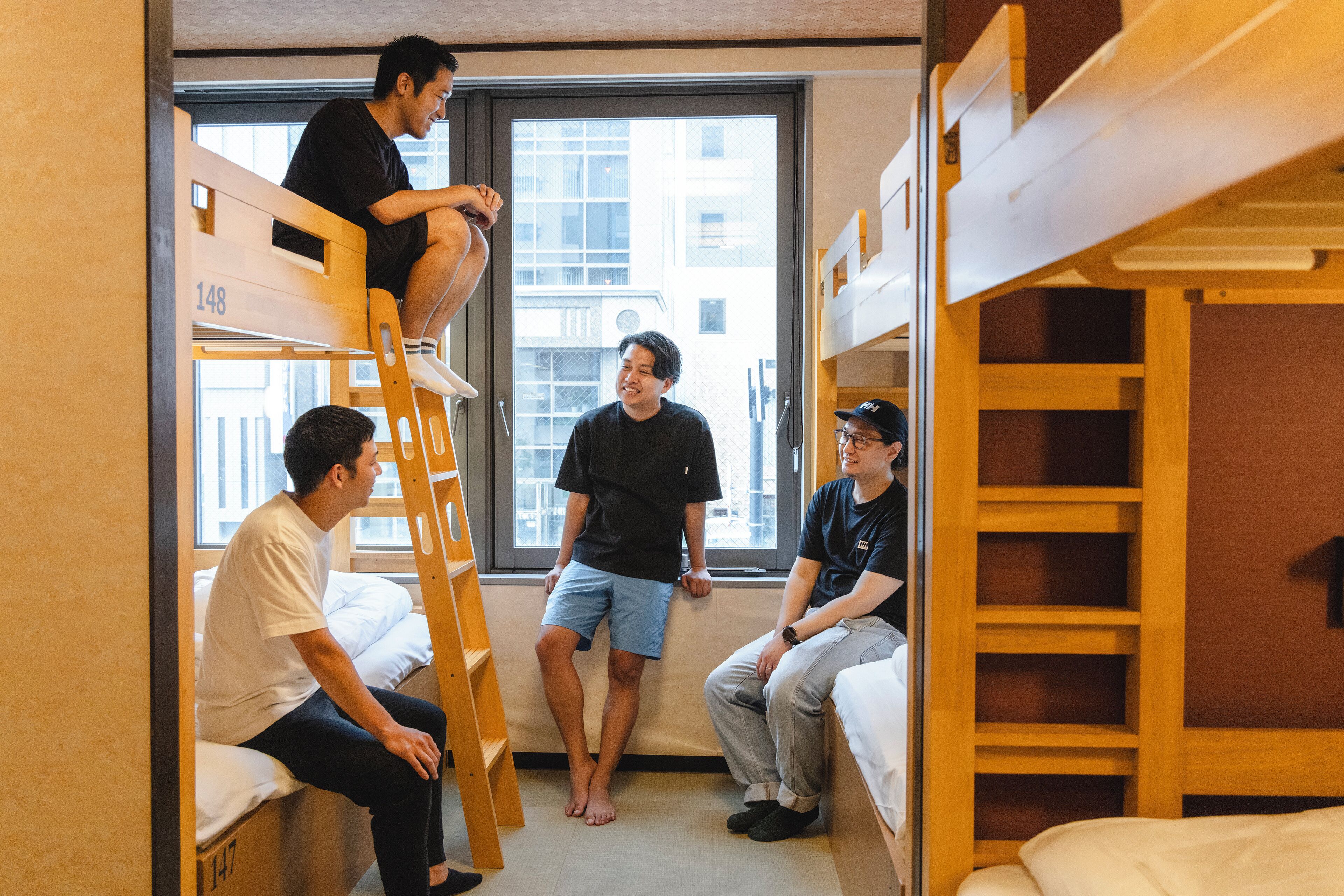 A dormitory room with bunk beds, a ladder, and a window with a view of buildings.