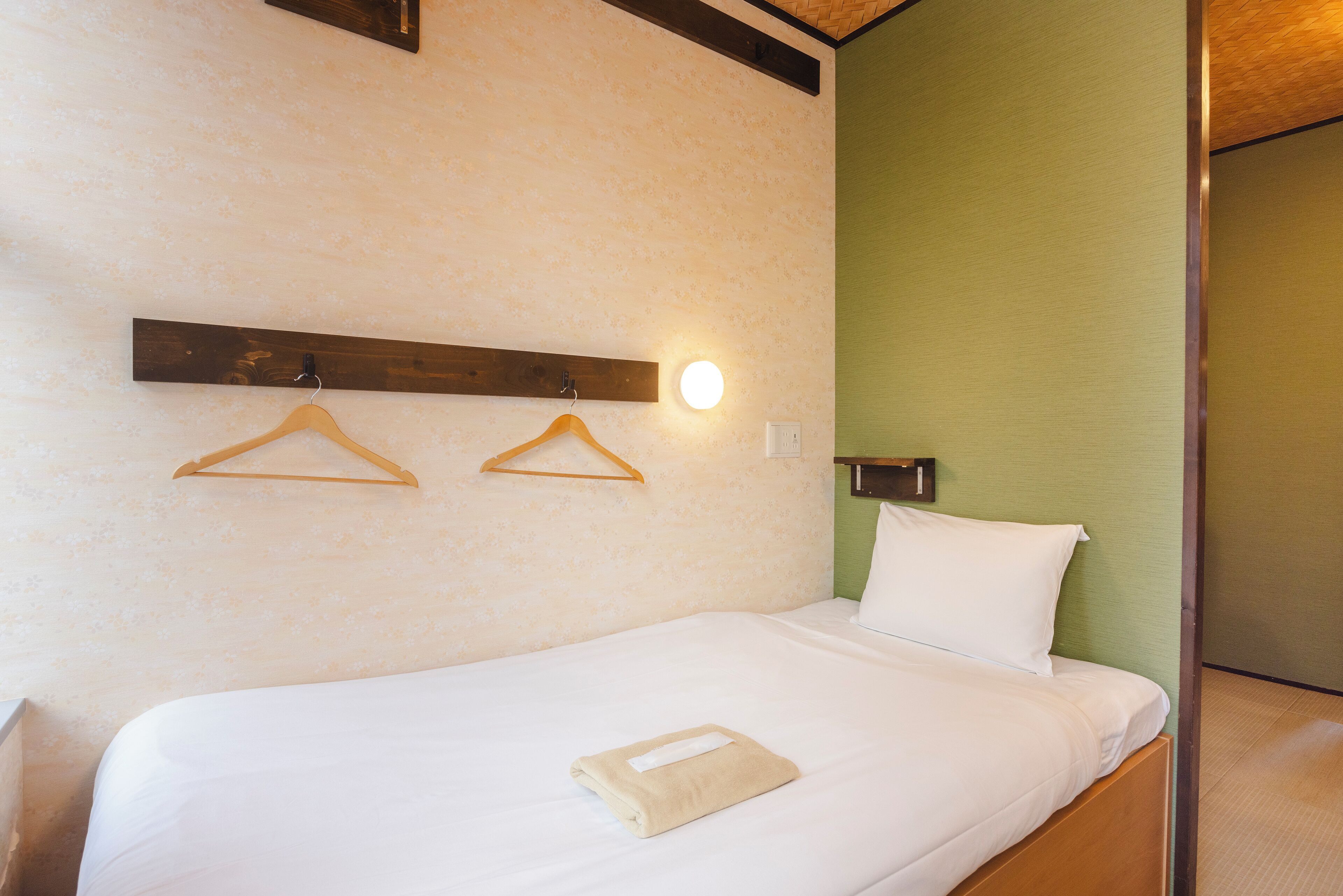 Una cama bien hecha con una almohada blanca y una toalla doblada, percheros de madera en la pared y un interruptor de luz.