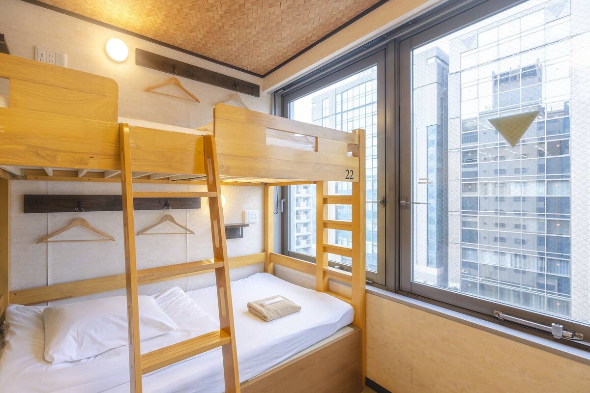 Une chambre avec des lits superposés en bois, une fenêtre donnant sur des immeubles et un luminaire suspendu.