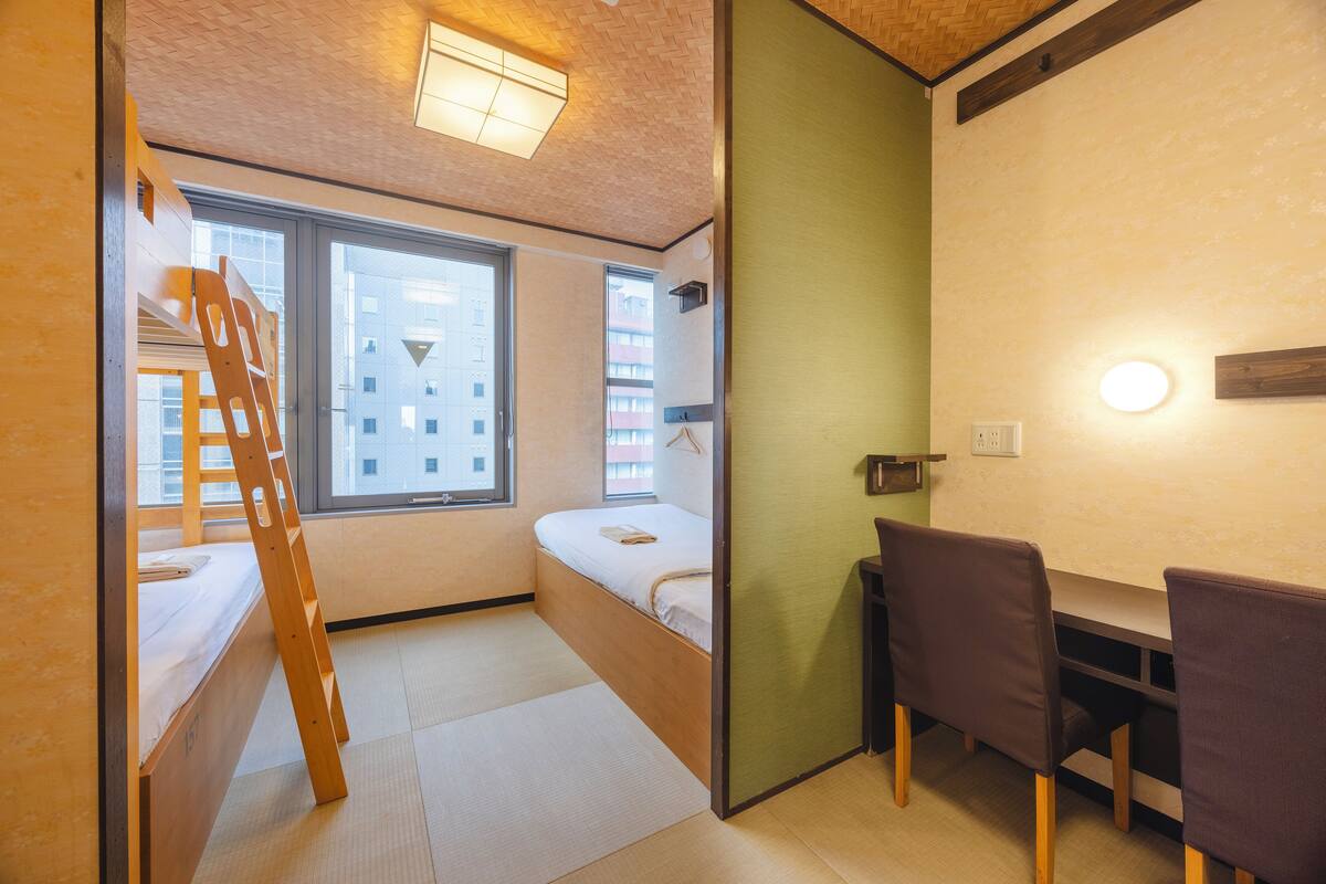 Une petite pièce comprenant un lit superposé, un bureau avec des chaises et une fenêtre donnant sur les bâtiments environnants.