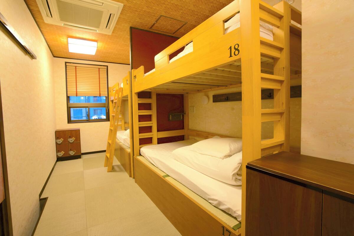 Une chambre de dortoir avec des lits superposés, numérotés 18 et 17, des meubles en bois, une fenêtre avec des stores et un petit placard.