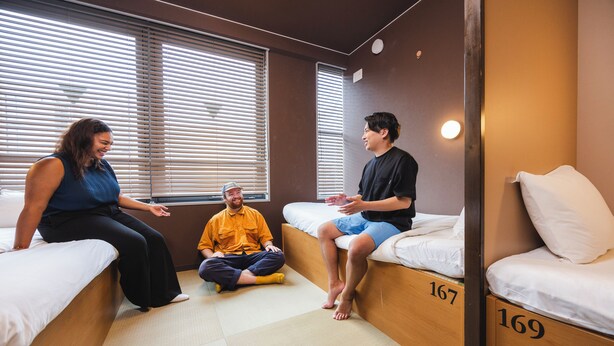 A room with three beds, each with a number. There are windows with blinds, a wooden floor, and a wall-mounted light. - Cogo Ryogoku (Tokyo)