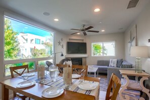 A modern living room with a dining area, large windows, a ceiling fan, and a flat-screen TV.