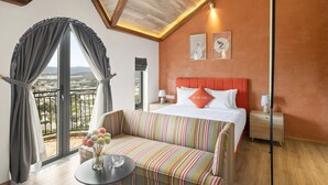 Habitación de hotel con un balcón grande, una cama con cabecera naranja, un sofá a rayas y una mesita de noche con lámpara.