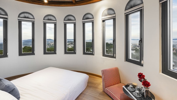 Un dormitorio con una cama grande, una silla y una mesa con un jarrón de flores. Hay varias ventanas con arcos y vistas a la ciudad.