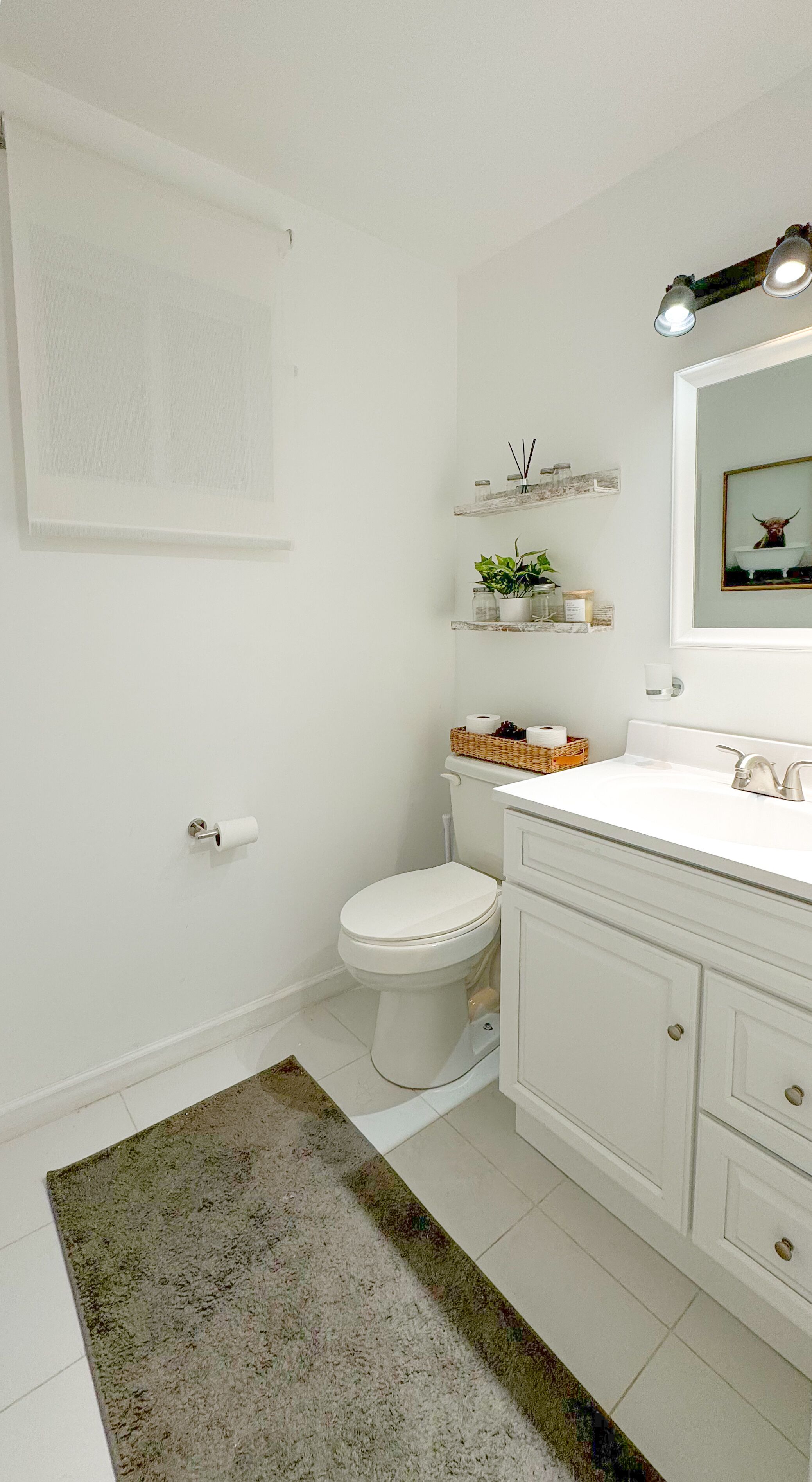 Une salle de bain comprenant des toilettes, un lavabo et un miroir. Des étagères se trouvent au-dessus du lavabo, où sont disposés des objets décoratifs.
