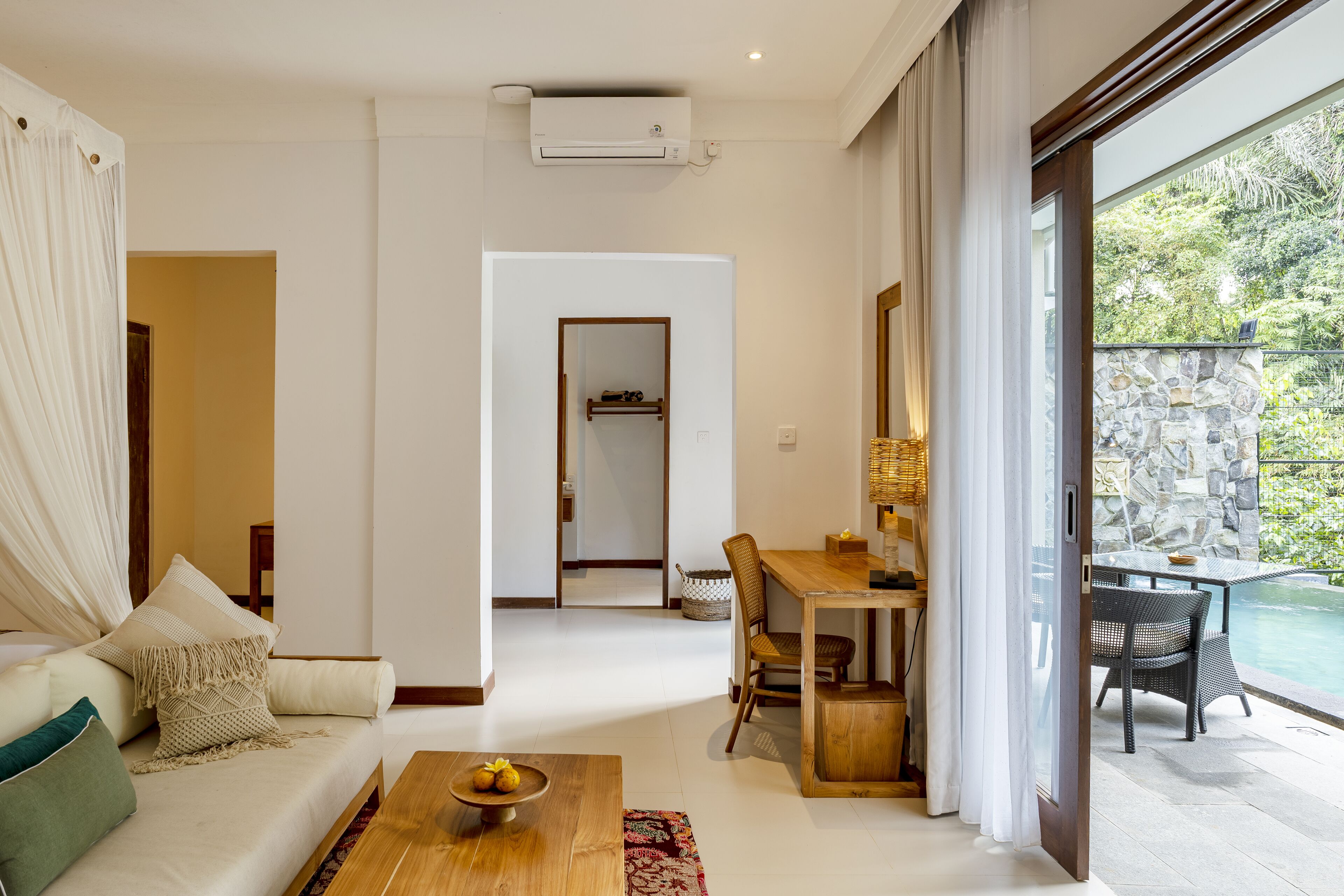 A modern living room with a sofa, coffee table, and dining area. There is a view of a pool and greenery outside.