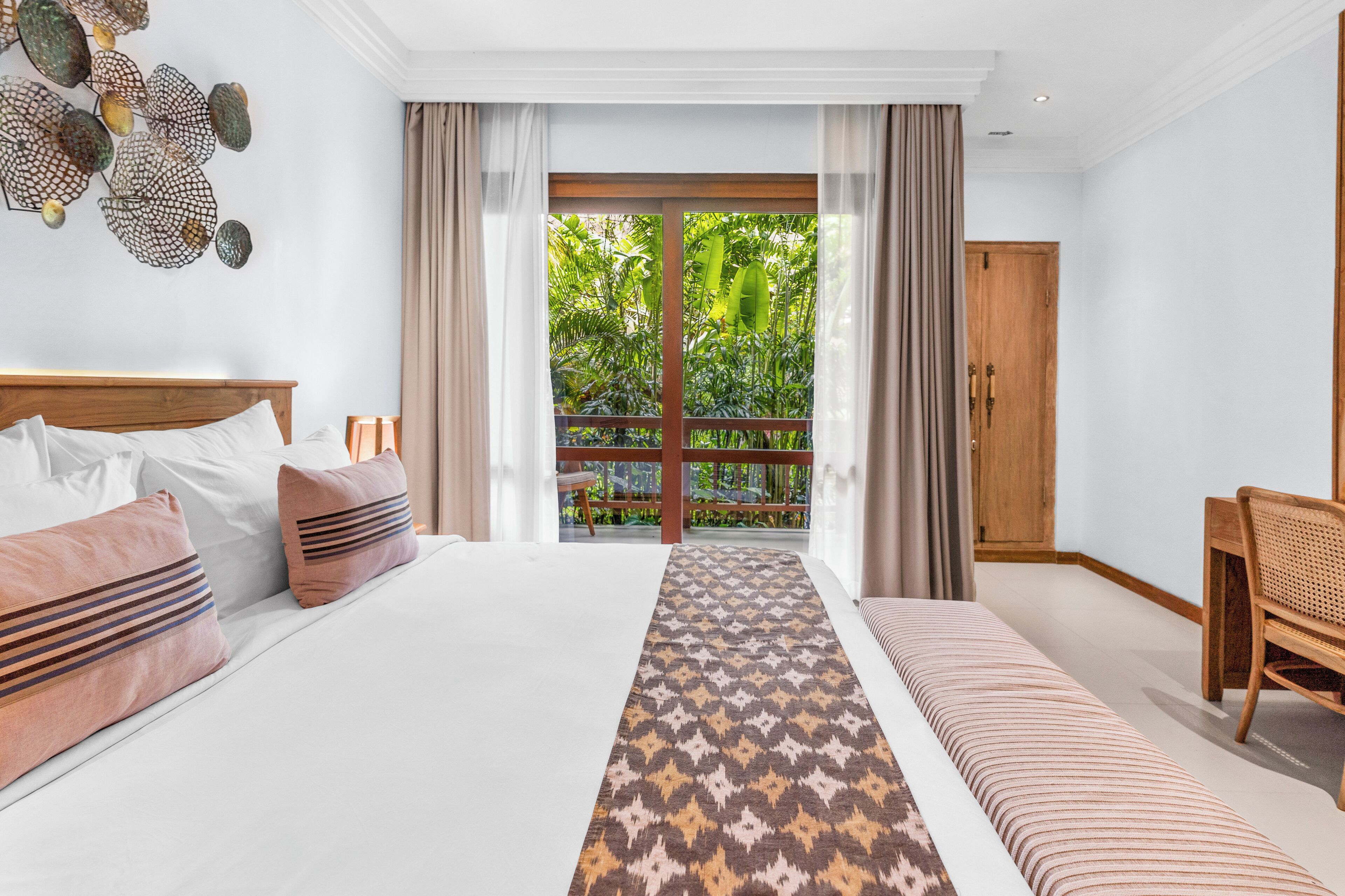 A bedroom with a large bed, a wooden chair, a patterned rug, and a view of greenery through a sliding door.