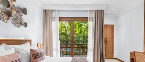 A bedroom with a large bed, a wooden chair, a patterned rug, and a view of greenery through a sliding door.
