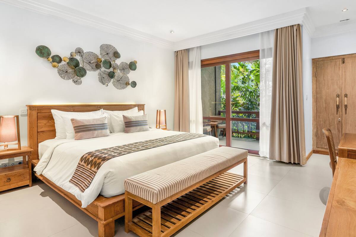 A modern bedroom with a large bed, a wooden bench, a nightstand, and a view of the outdoors through a sliding glass door.