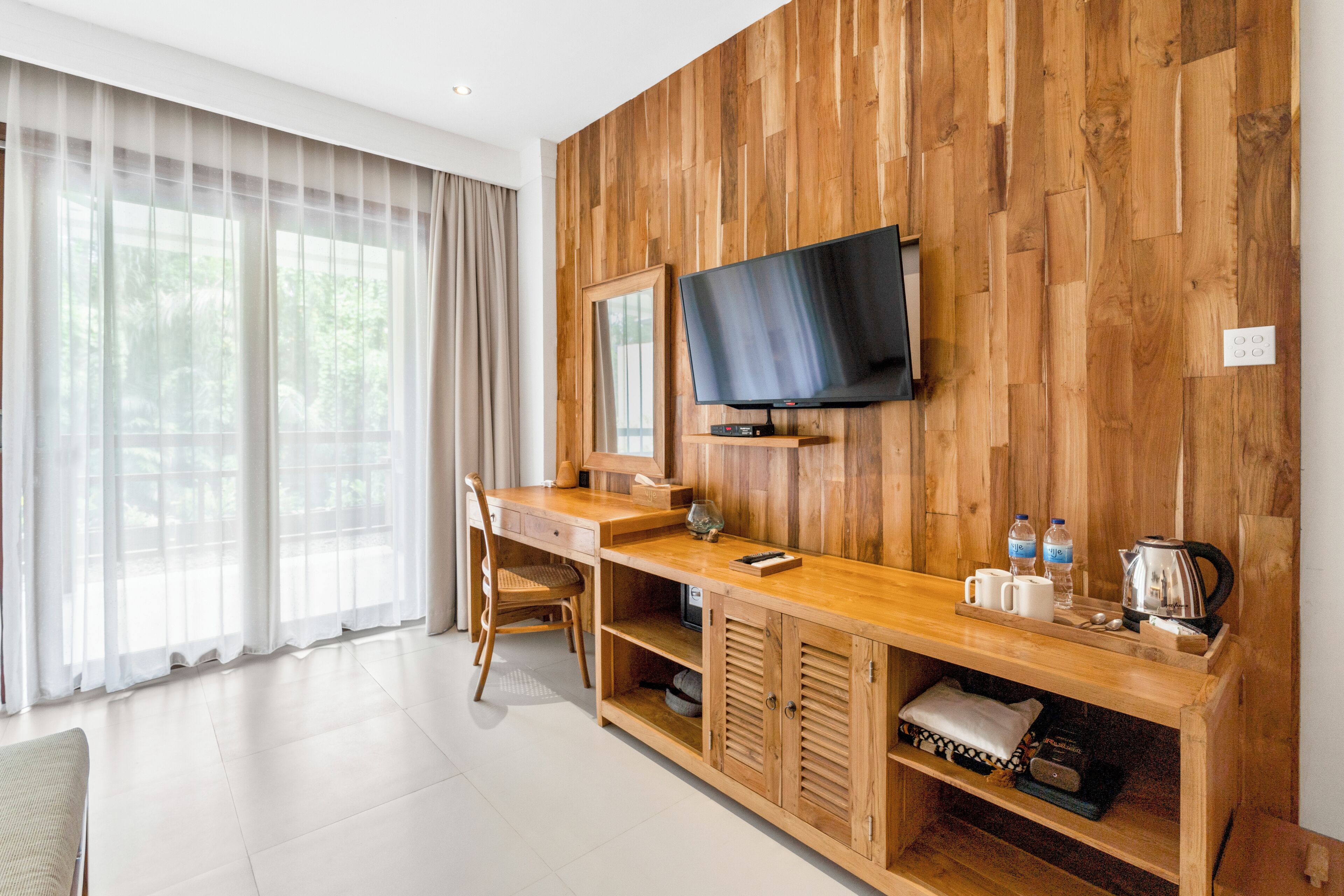 A modern hotel room with a wooden wall, a flat-screen TV, a wooden desk, a chair, and a large window with sheer curtains.