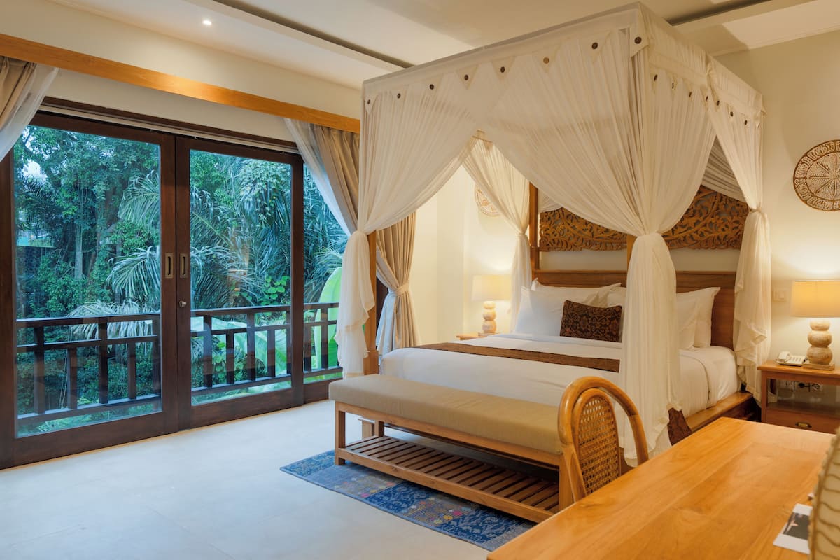 A bedroom with a four-poster bed, a wooden bench, a table, and a view of lush greenery through large sliding glass doors.