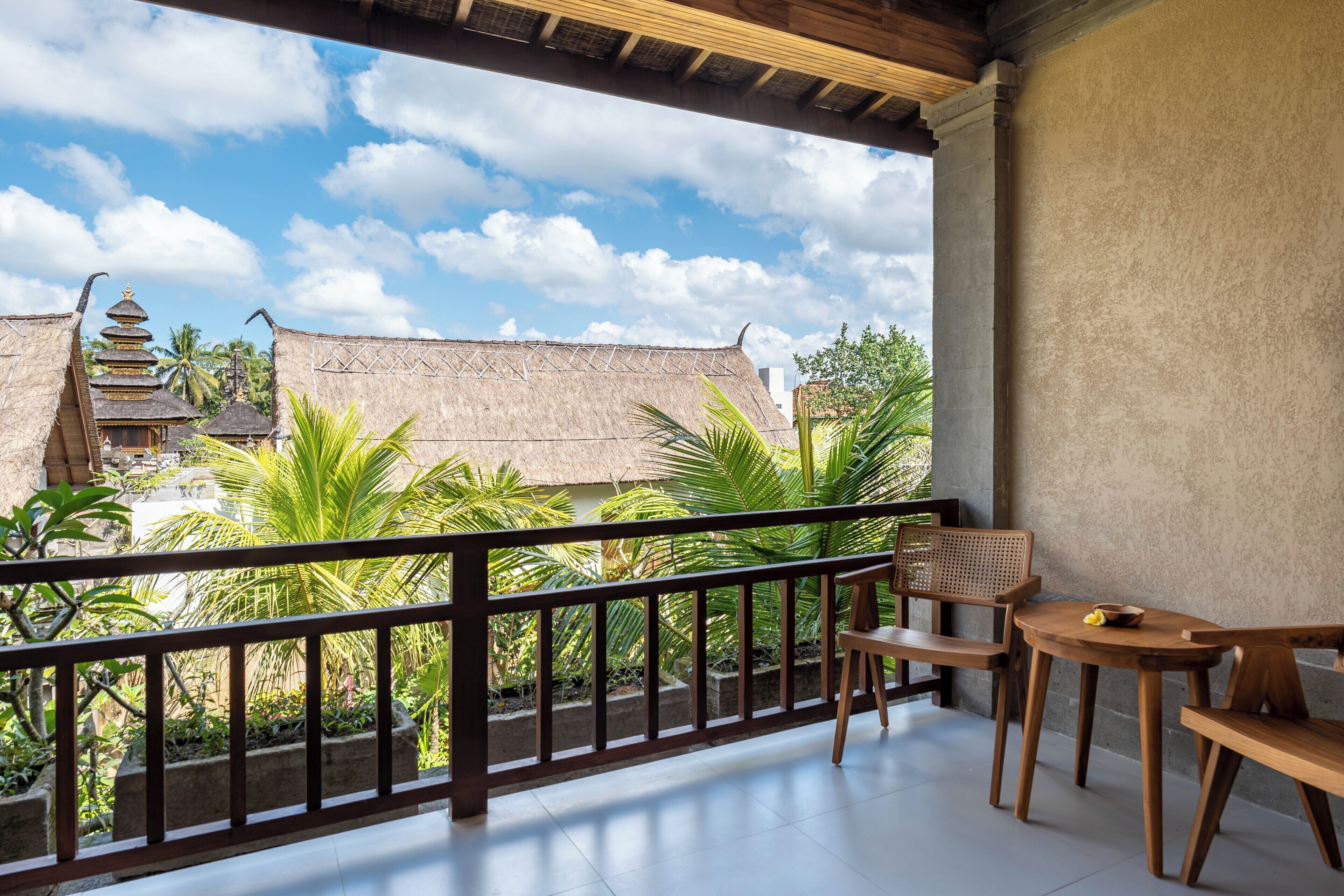 A balcony with a wooden table and chairs, overlooking a traditional building with a thatched roof and a pagoda-like structure.