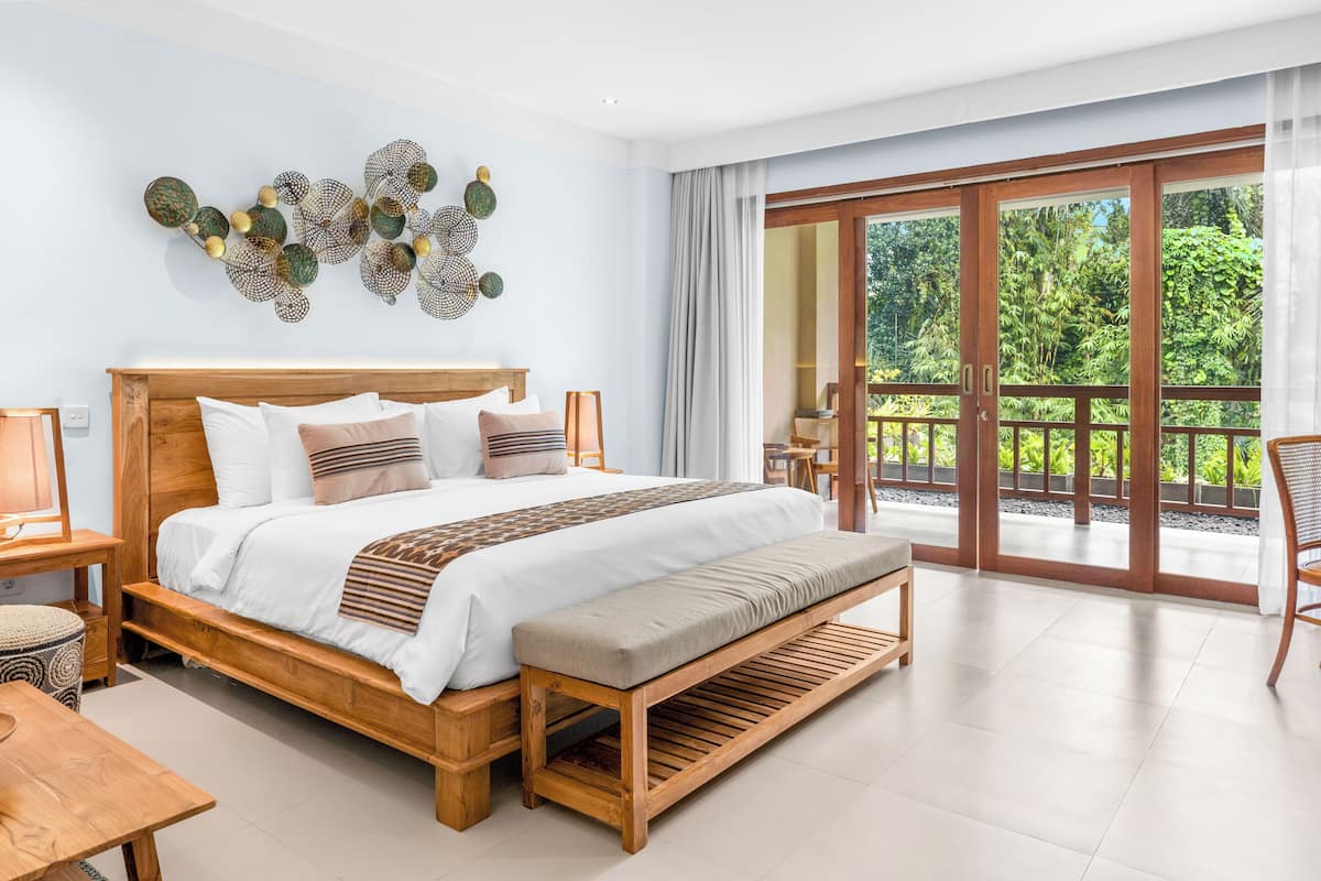 A spacious bedroom with a large bed, a wooden bench, and a view of lush greenery through a sliding glass door.