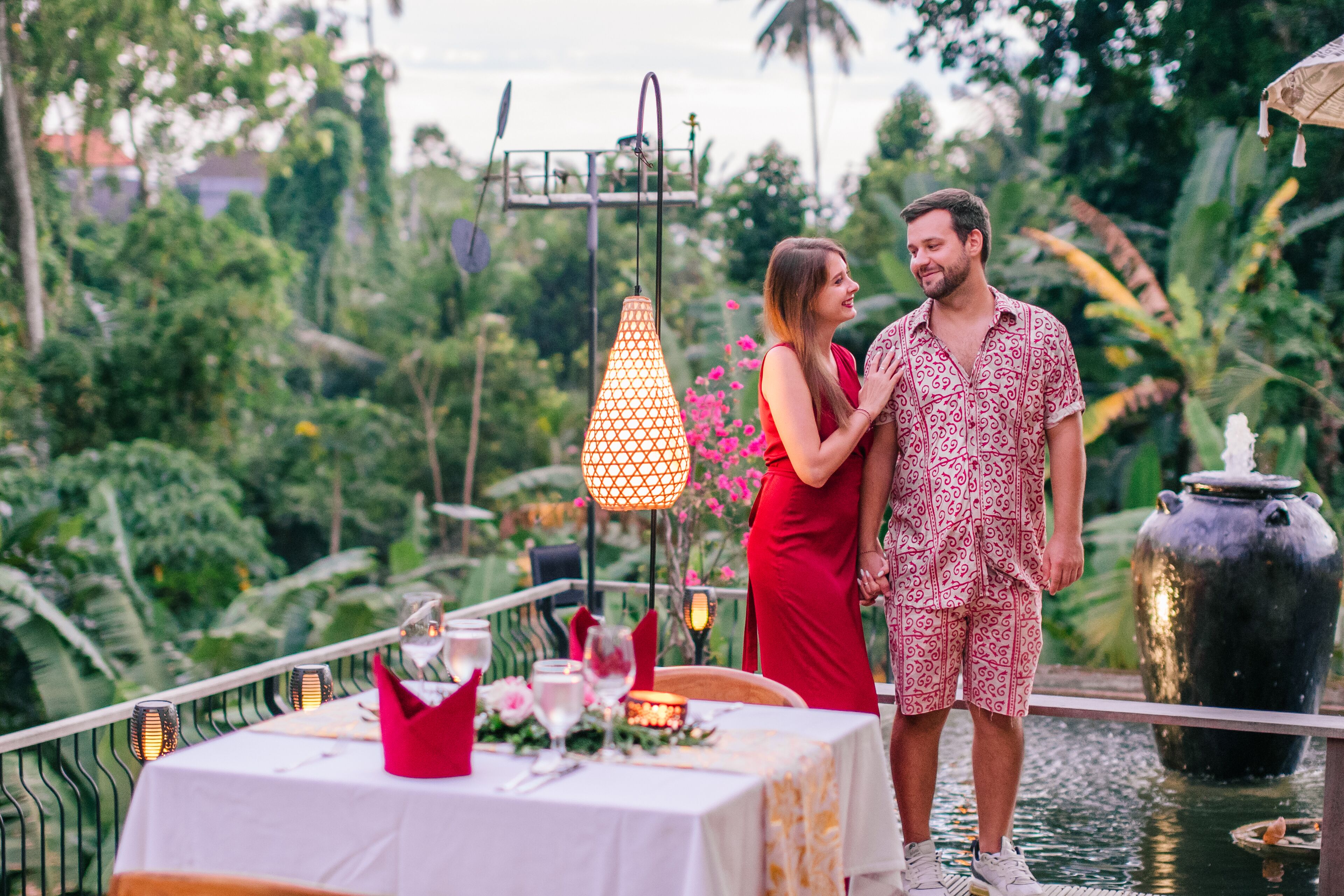 A couple standing by a table set for a meal, with a decorative lamp and a water feature in the background.
