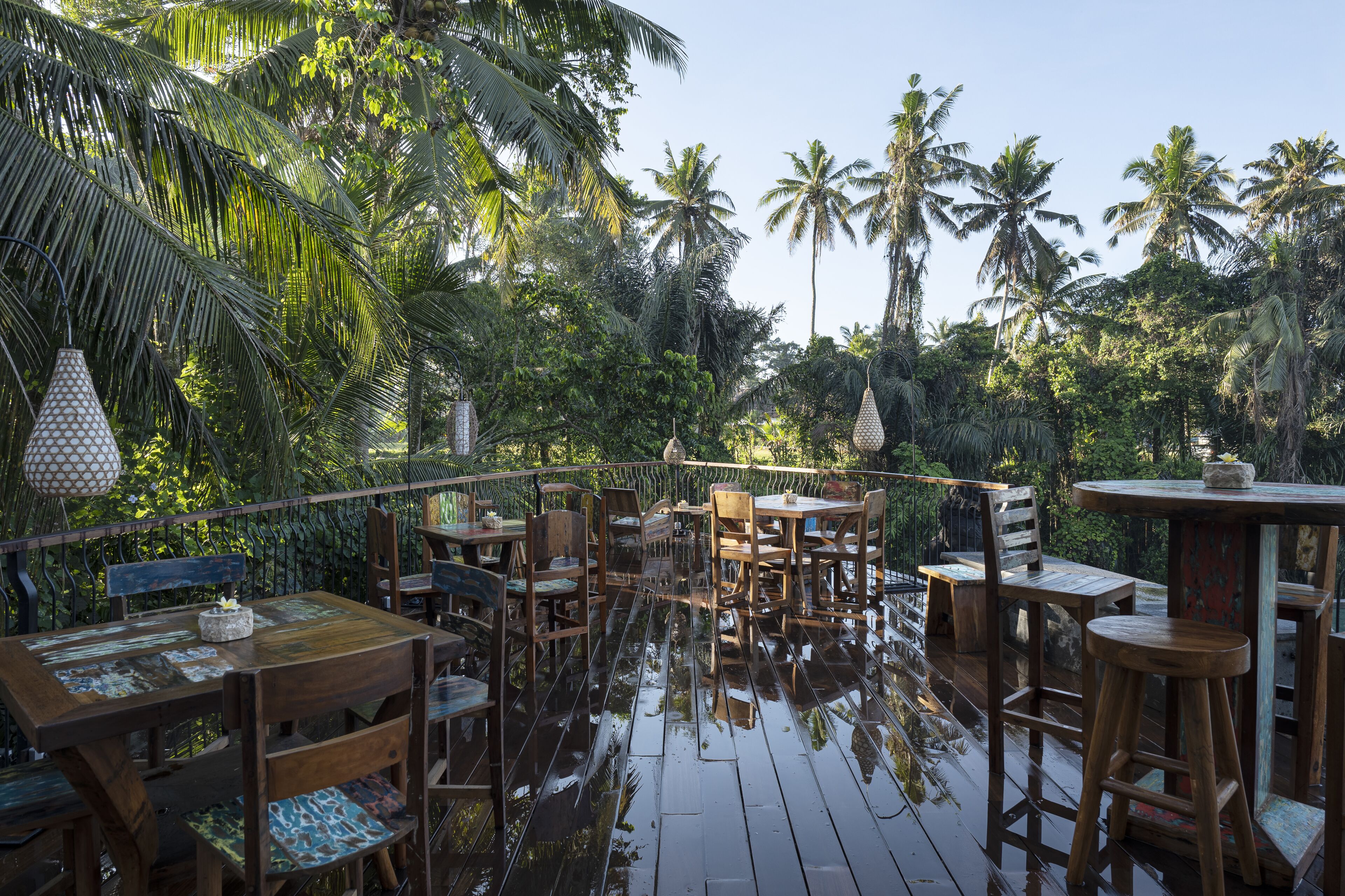 An outdoor wooden deck with tables and chairs, surrounded by lush tropical vegetation and palm trees.