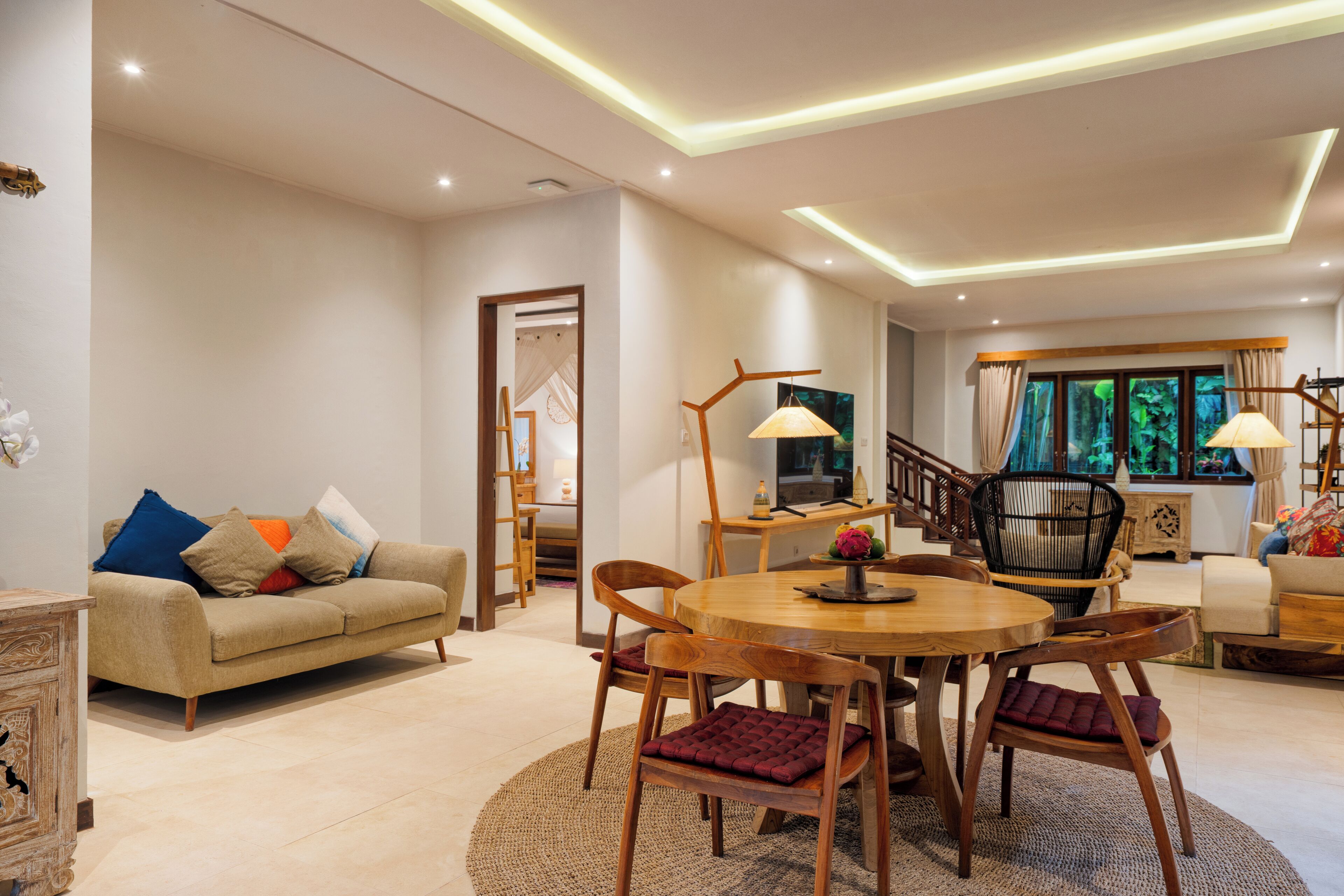 A spacious living room with a round wooden dining table, cushioned chairs, a sofa with colorful pillows, and a staircase leading to another level.