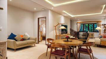 A spacious living room with a round wooden dining table, cushioned chairs, a sofa with colorful pillows, and a staircase leading to another level.