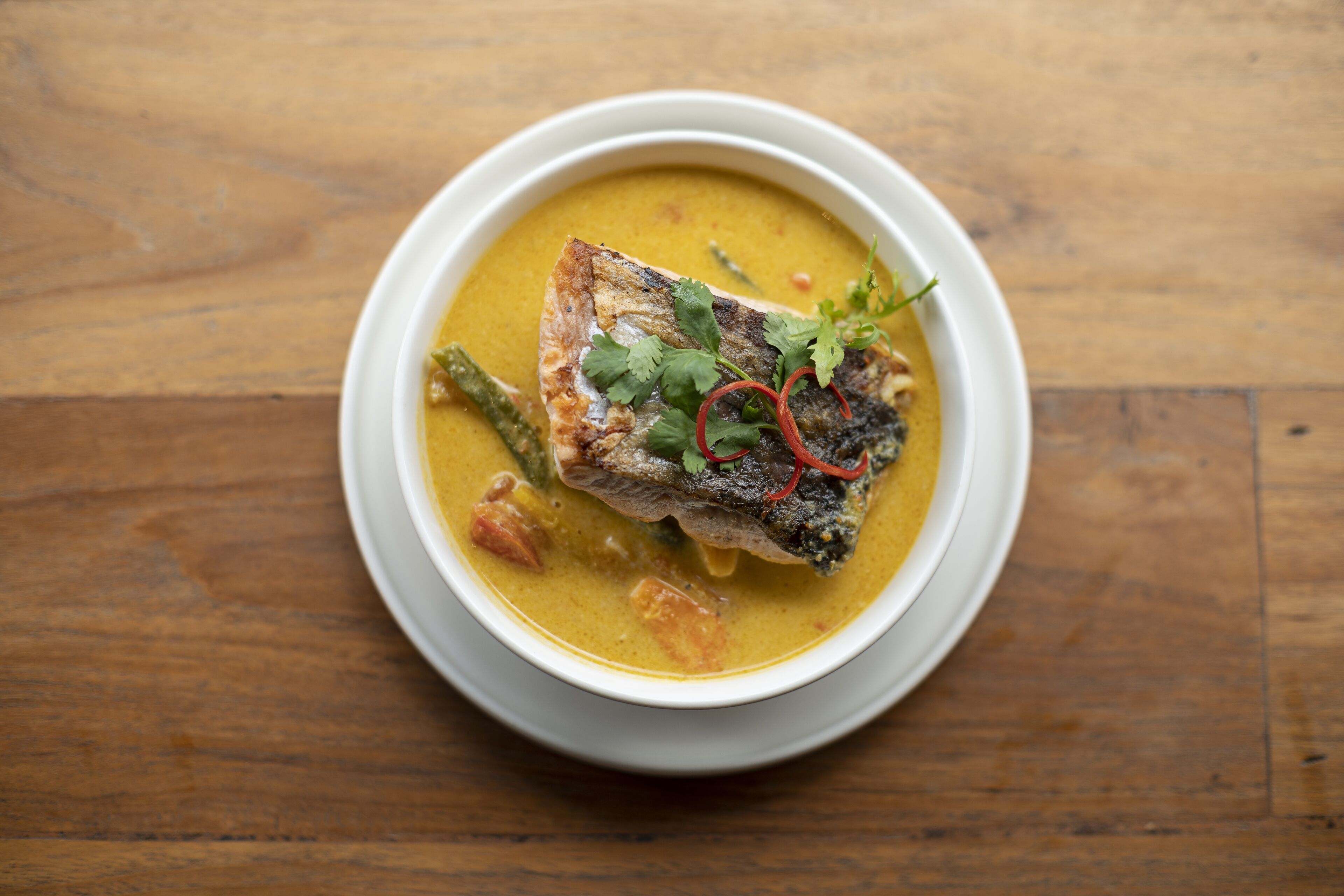 A bowl of fish soup with a piece of fish and vegetables on a wooden table.
