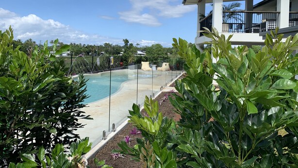 A swimming pool with a glass barrier, surrounded by lush greenery and a modern building with a balcony.