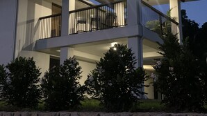 A modern two-story house with a balcony, stone wall, and surrounding greenery.