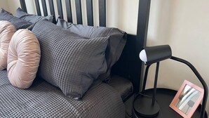 A neatly made bed with a grey textured duvet and several pillows, a black bedside table with a lamp and a picture frame, and a striped headboard. - Peaceful Coombs Apartment for Families (Coombs)