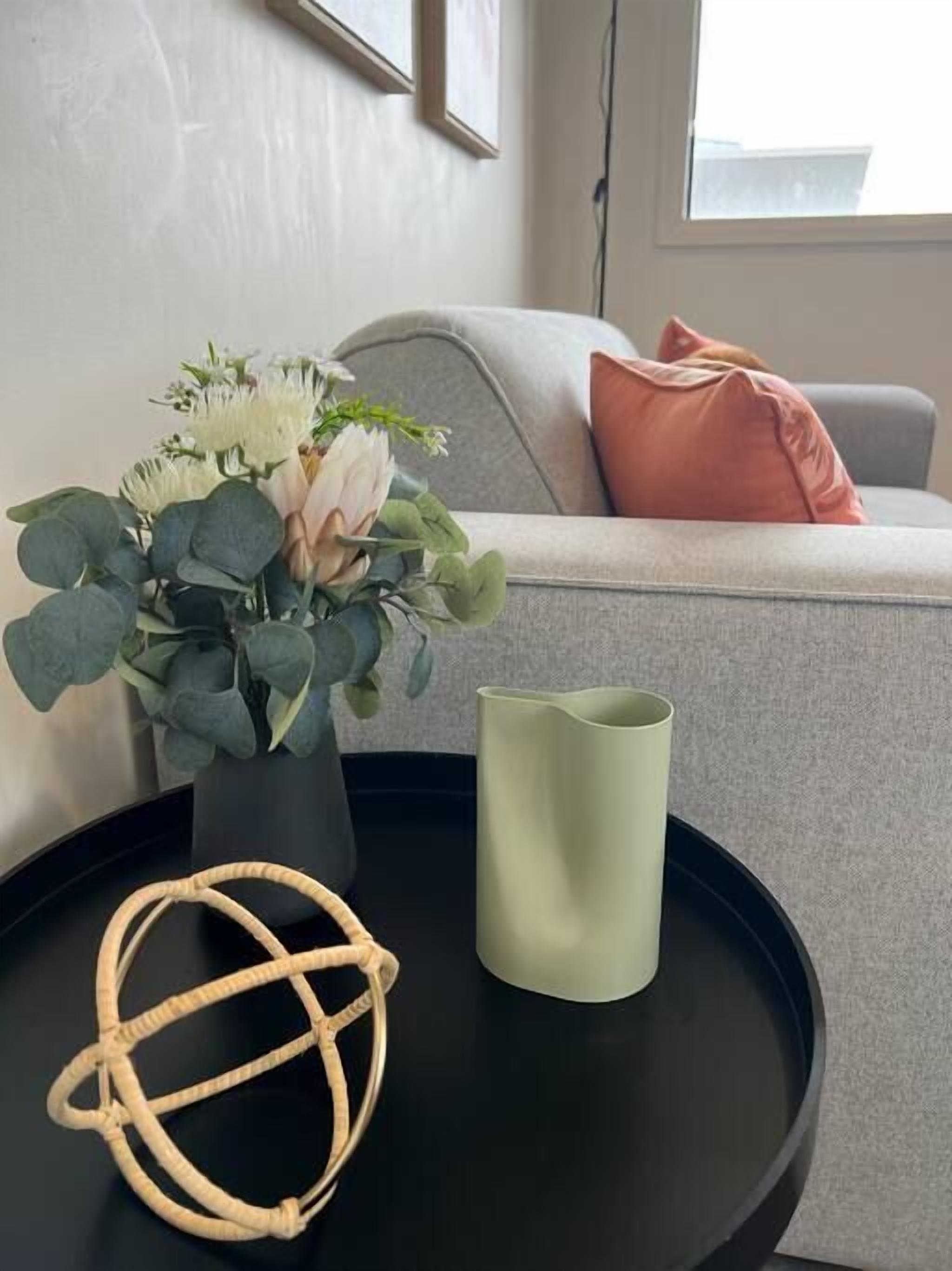 A living room with a grey sofa, a pink pillow, a vase with flowers, a green candle, and a woven object on a black table.