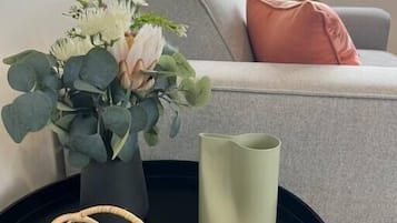 A living room with a grey sofa, a pink pillow, a vase with flowers, a green candle, and a woven object on a black table.