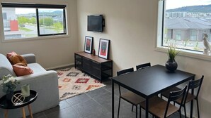 A modern living room with a grey sofa, a small coffee table, a TV mounted on the wall, and a dining area with a table and chairs. - Peaceful Coombs Apartment for Families (Coombs)