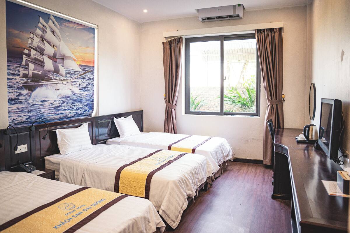 A hotel room with two beds, a large window with curtains, a painting of a ship on the wall, and a desk with a television.