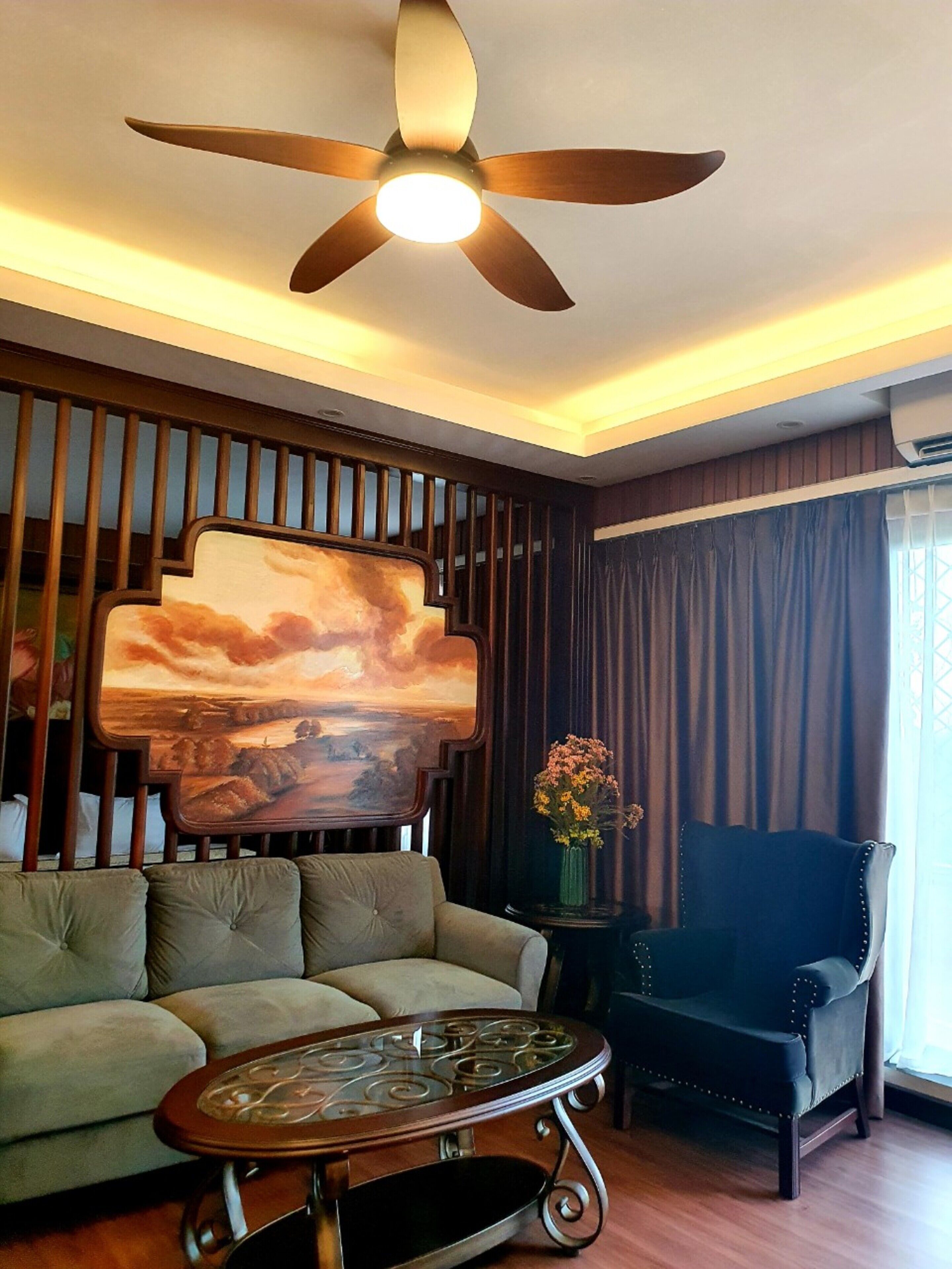 A living room with a sofa, a chair, a coffee table, a ceiling fan, and a large painting on the wall.