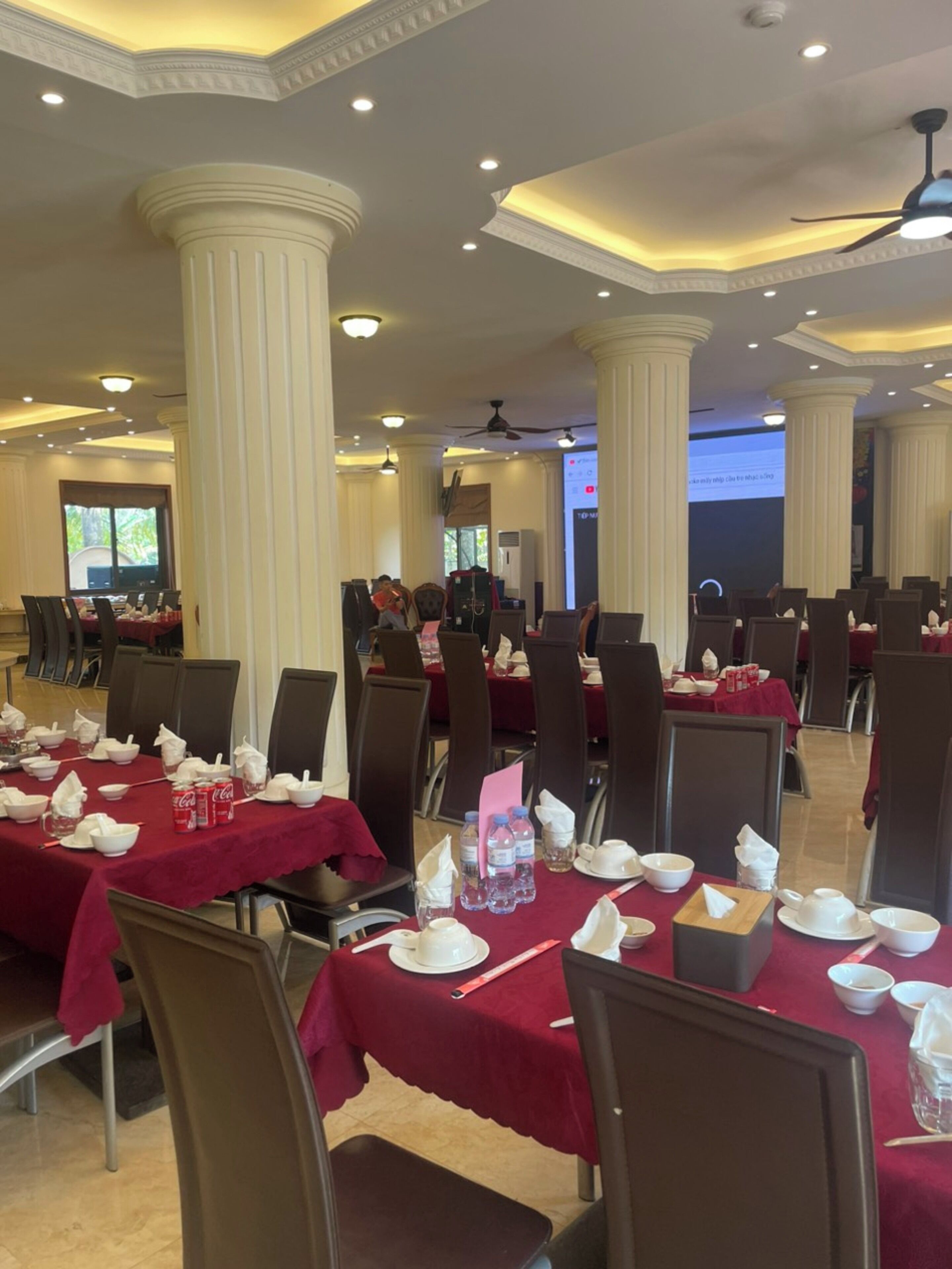 A banquet hall with tables set for a formal event, featuring white columns, recessed lighting, and a projection screen in the background.