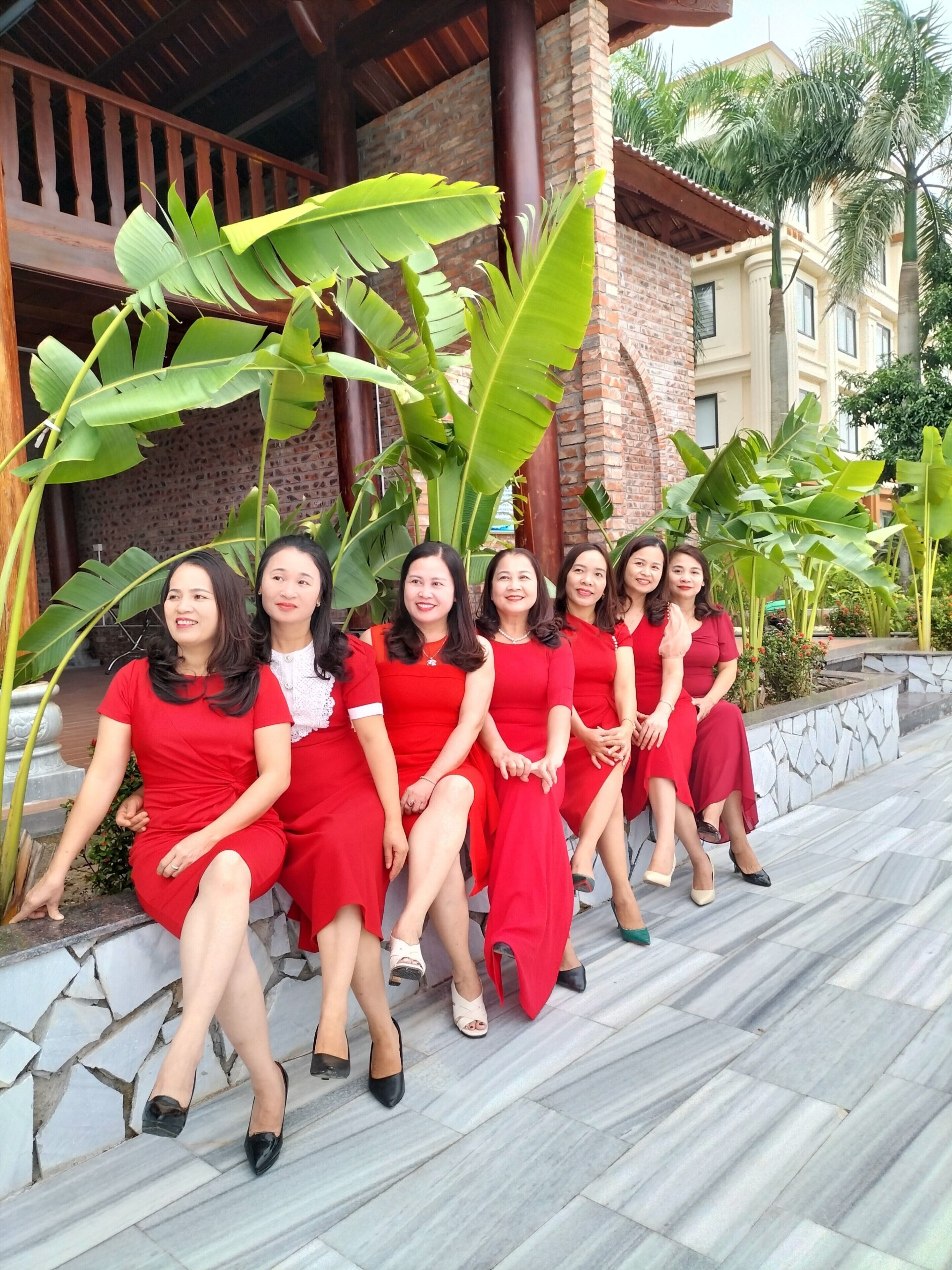 A group of women in red dresses seated on stone steps under a wooden pergola with large banana leaves.