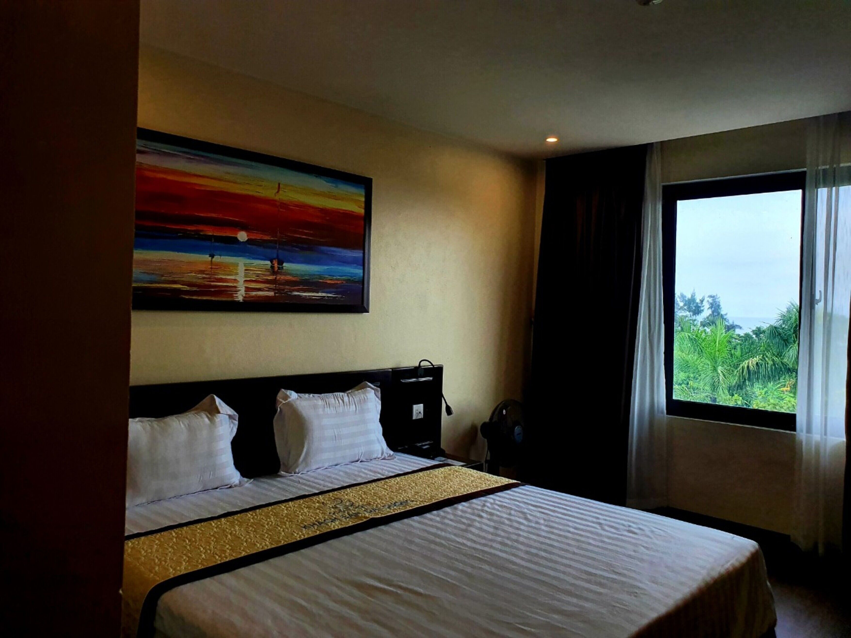 Une chambre d’hôtel avec un grand lit, un tableau représentant un coucher de soleil, une fenêtre donnant sur un paysage verdoyant et une table de chevet avec une lampe.