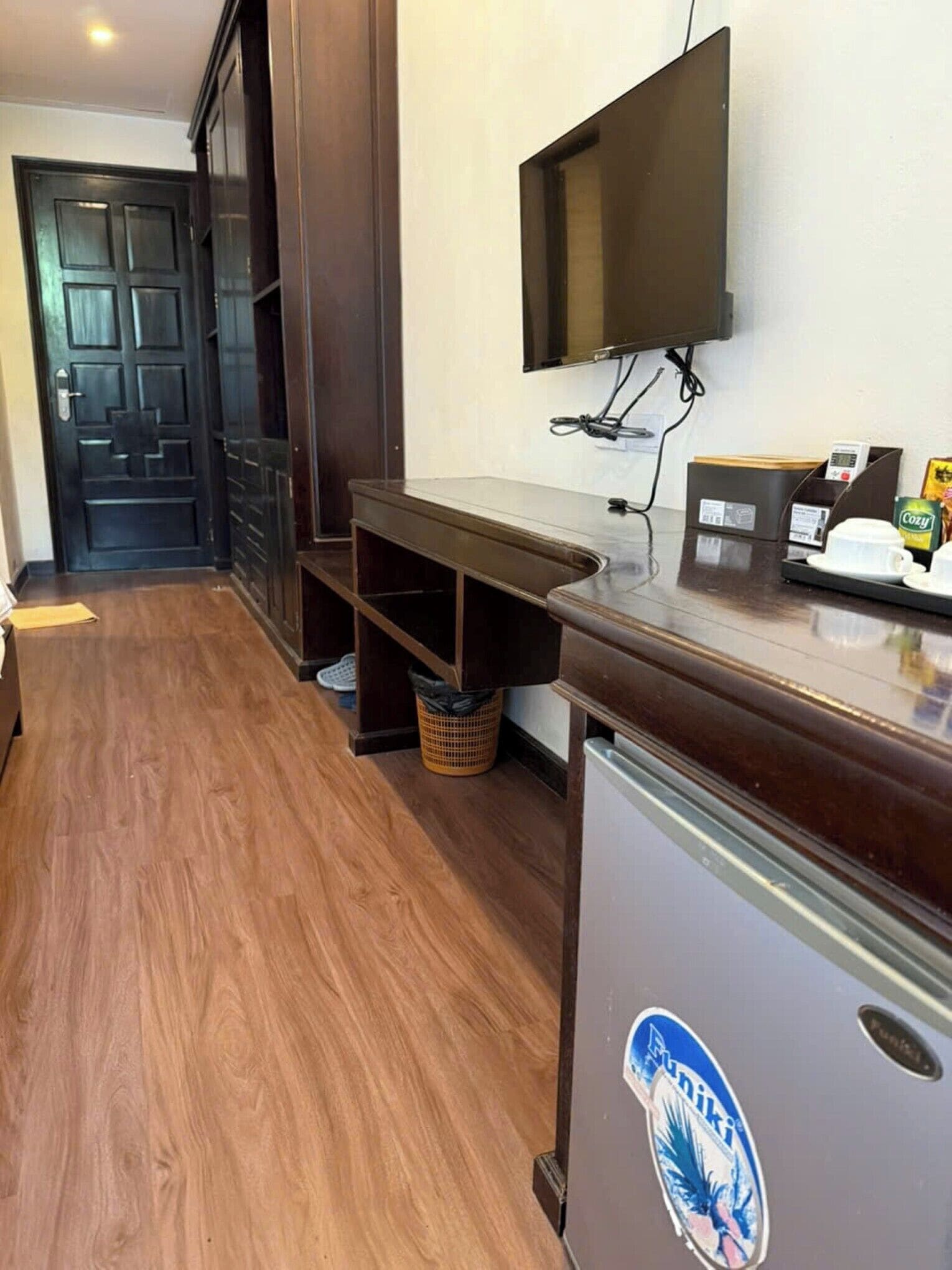 A hotel reception area with a wooden floor, a dark reception desk, a mounted TV, and a trash bin.