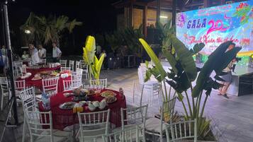 Un área de comedor al aire libre con mesas dispuestas para una comida, rodeada de sillas y grandes plantas tropicales.