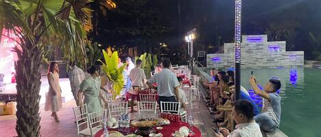 An outdoor dining area with round tables covered in red tablecloths, surrounded by white chairs.