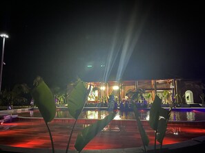 Una vista nocturna de un área de piscina con escenario y asientos, rodeada de palmeras e iluminada por focos.