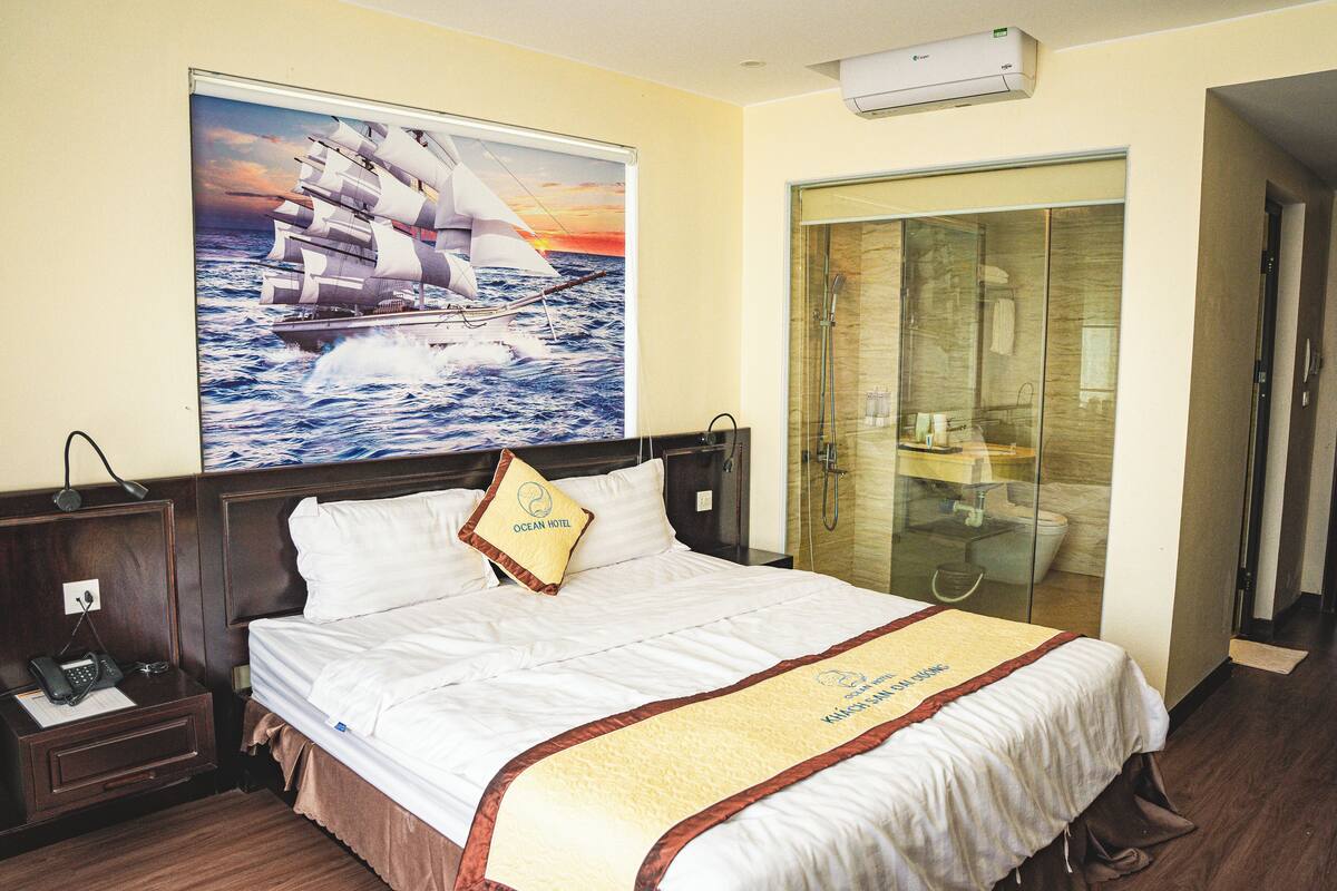 A hotel room with a bed, a nightstand with a phone, a glass-enclosed shower, and a painting of a sailboat on the wall.