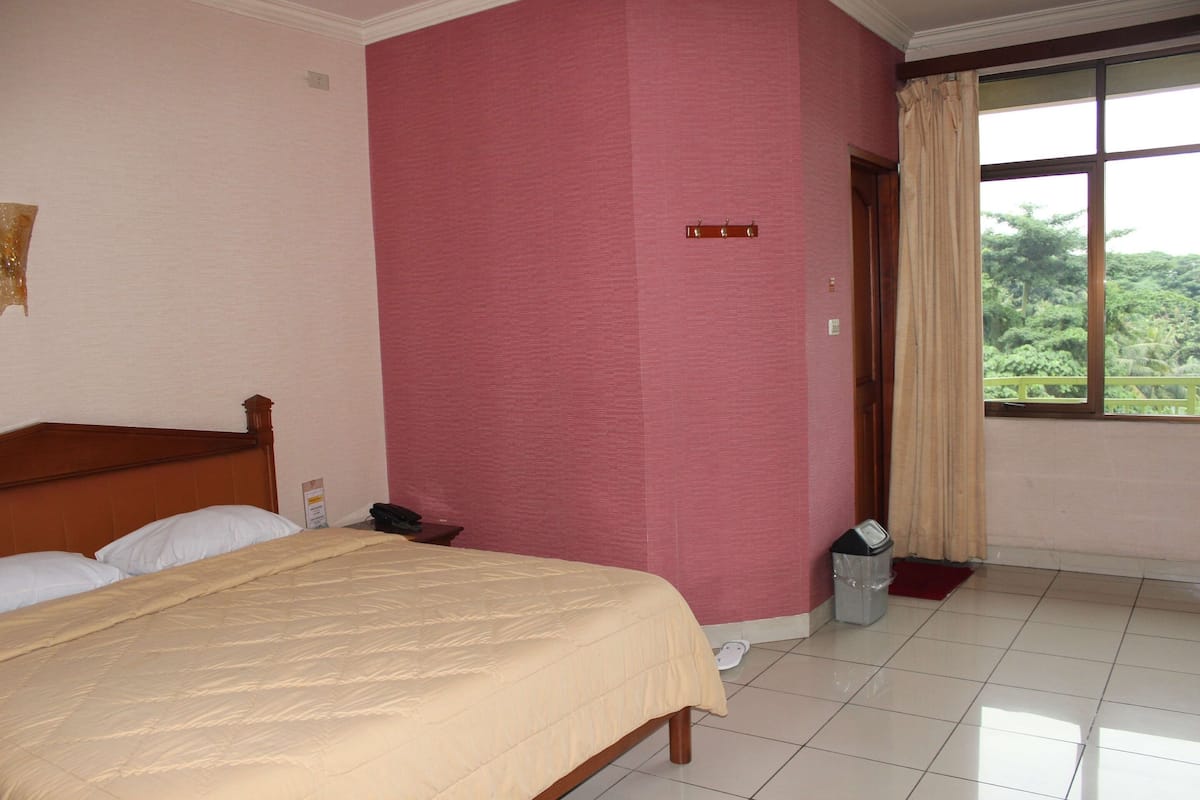 A hotel room with a bed, a window with curtains, a trash bin, and a door.