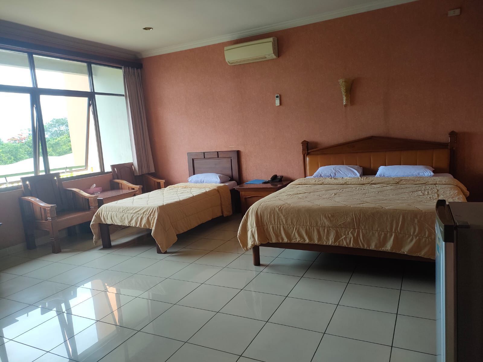 A hotel room with two beds, a desk, a chair, a window with curtains, and an air conditioning unit.